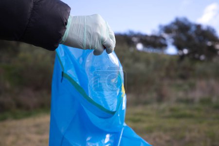 Detail of a volunteer's hand picking up plastic bottle from the forest and putting in the plastic bag. Concept of Earth Day and World Environment Day June 5.