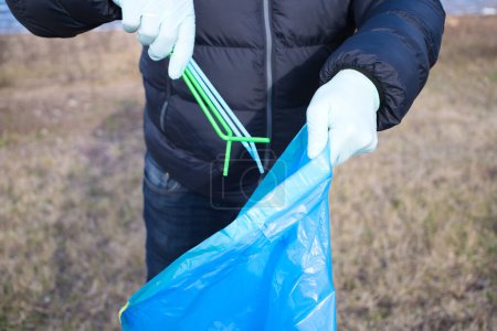 Detail of a volunteer's hand picking up plastic straws from the forest and putting them in the plastic bag. Concept of Earth Day and World Environment Day June 5.