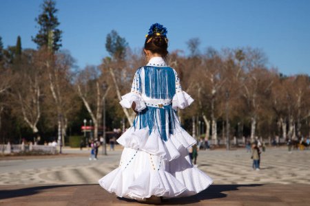 Foto de Una niña bailando flamenco vestida con un vestido blanco con volantes y flecos azules en una famosa plaza de Sevilla, España. La chica tiene flores en la cabeza. Flamenco, patrimonio cultural de la humanidad - Imagen libre de derechos