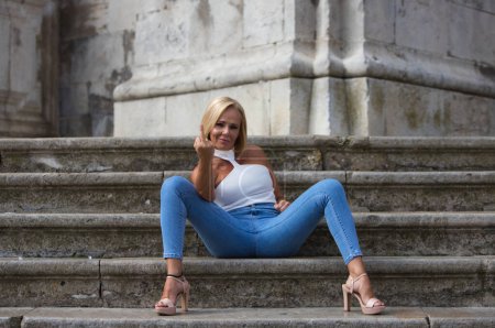 Pretty blonde adult woman in jeans and white top sits on the stairs and makes a rude and offensive gesture with her finger. Concept fuck you.
