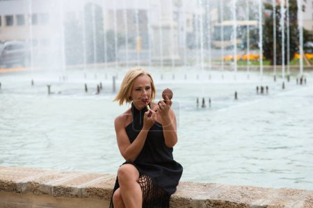 Beautiful blonde mature woman is dressed in a fringed black dress and is sitting on the edge of a fountain. The woman paints her lips and looks into the small mirror