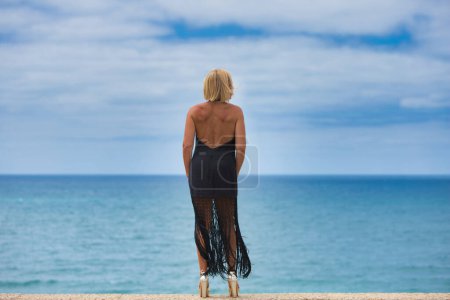 Reife, blonde, schöne Frau in elegantem schwarzen Kleid blickt sehnsüchtig auf den Horizont über dem Atlantik in cadiz, Spanien. Die Frau ist traurig und nostalgisch. Foto von hinten aufgenommen