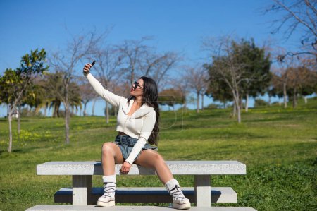 Junge und schöne brünette Spanierin mit Sonnenbrille auf einer Parkbank sitzend. Das Mädchen ist lässig gekleidet und macht ein Selfie mit ihrem Handy, während sie den sonnigen Frühlingstag genießt