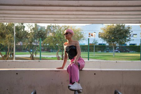 Photo for Young gay boy with pink hair and make-up and sunglasses is sitting on a wall resting, in the background a park with trees. Concept of equality and lgbtq rights - Royalty Free Image