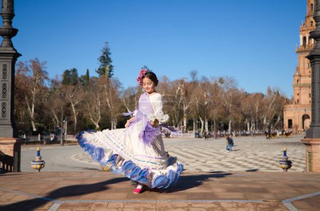 Foto de Una niña bailando flamenco vestida con un vestido beige con volantes y flecos morados en una famosa plaza de Sevilla, España. La chica tiene flores en la cabeza y el pelo en un moño. - Imagen libre de derechos