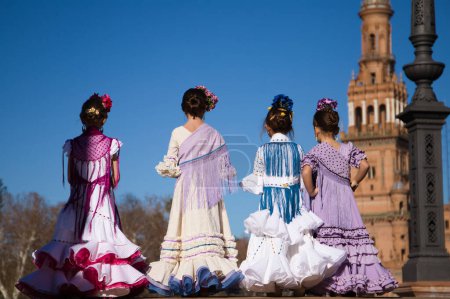 Foto de Cuatro niñas bailando flamenco vestidas con el típico traje de flamenca posan para una foto en una famosa plaza de Sevilla, España. Las chicas tienen flores en la cabeza. Al fondo una antigua torre cuadrada - Imagen libre de derechos