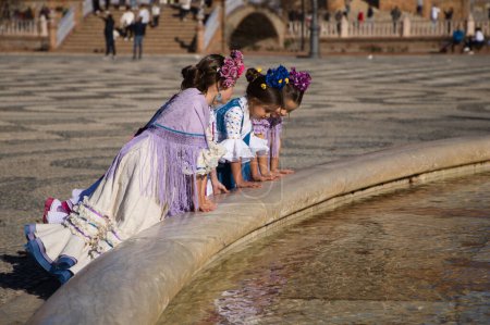 Foto de Cuatro niñas bailando flamenco vestidas con el típico traje de flamenca que asoman a la fuente en una famosa plaza de Sevilla, España. En el fondo los arcos y columnas de la famosa plaza - Imagen libre de derechos
