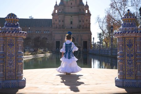 Una chica guapa bailando flamenco con un vestido de volantes y flecos en una famosa plaza de Sevilla, España. La chica lleva una flor en el pelo y le da la espalda. Al fondo una torre