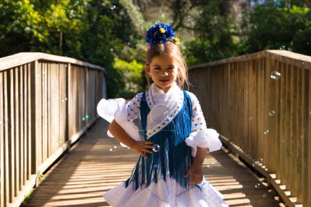 Foto de Una bonita niña bailando flamenco con un típico vestido gitano con volantes y flecos pasea sobre un puente de madera en un famoso parque de Sevilla, España. La chica posa entre las burbujas de jabón - Imagen libre de derechos