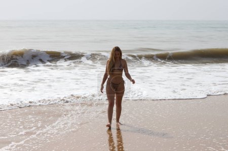 beautiful young blonde woman coming out of the sea is walking along the shore of the beach. The woman is wearing a leopard bikini and enjoying her summer holidays in spain. Travel concept