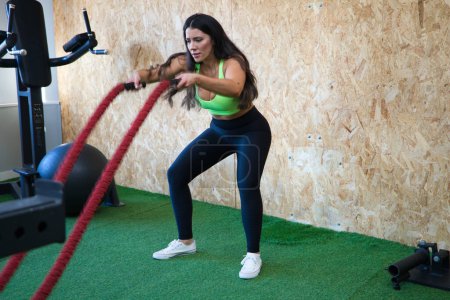 Mujer morena joven y hermosa está haciendo ejercicios de crossfit con cuerdas, se concentra y ejerce fuerza con los brazos y las manos. Concepto salud y deporte
