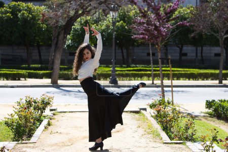Beautiful woman with long curly hair, dancing flamenco artfully in a park in Seville, Spain. Dressed in long black skirt and white shirt. Flamenco, cultural heritage of humanity