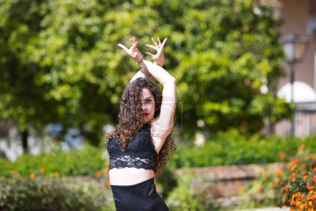 Beautiful woman with long curly hair, dancing flamenco artfully in a park in Seville, Spain. Dressed in long black skirt and black top. Flamenco, cultural heritage of humanity