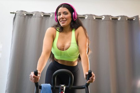 Young and beautiful brunette woman training on the bike in the gym. The girl is listening to music on her headphones. She is dressed in a top and bikini bottoms. Concept of health and sport