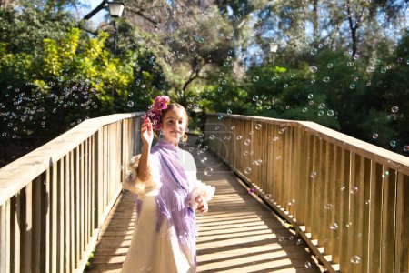 Una bonita niña bailando flamenco con un típico vestido gitano con volantes y flecos pasea sobre un puente de madera en un famoso parque de Sevilla, España. La chica juega con burbujas de jabón.