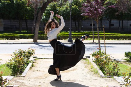 Beautiful woman with long curly hair, dancing flamenco artfully in a park in Seville, Spain. Dressed in long black skirt and white shirt. Flamenco, cultural heritage of humanity