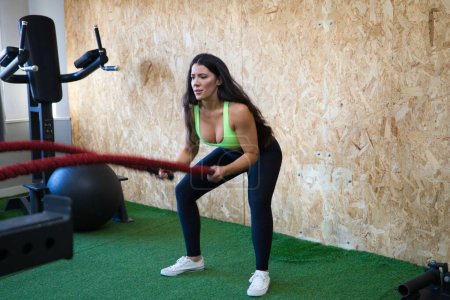 Jeune et belle femme brune fait des exercices de crossfit avec des cordes, elle est concentrée et exerce de la force avec les bras et les mains. Santé et sport concept