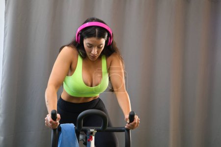 Young and beautiful brunette woman training on the bike in the gym. The girl is listening to music on her headphones. She is dressed in a top and bikini bottoms. Concept of health and sport