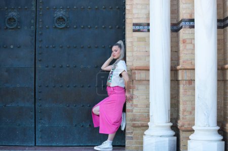Schöne junge blonde Frau lehnt am Türpfosten einer großen Tür eines monumentalen Gebäudes in der Stadt Sevilla. Das Mädchen stützt sich auf ein Bein und berührt ihren Kopf. Das Mädchen ist lässig gekleidet