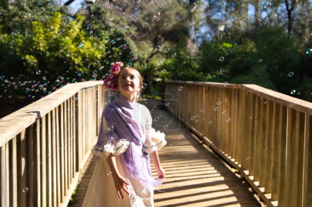 Foto de Una bonita niña bailando flamenco con un típico vestido gitano con volantes y flecos pasea sobre un puente de madera en un famoso parque de Sevilla, España. La chica juega con burbujas de jabón. - Imagen libre de derechos