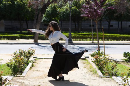 Foto de Hermosa mujer de pelo rizado largo, bailando flamenco artísticamente en un parque de Sevilla, España. Vestido con falda larga negra y camisa blanca. Flamenco, patrimonio cultural de la humanidad - Imagen libre de derechos