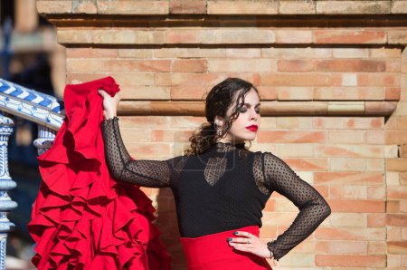 Schöne Frau tanzt Flamenco auf einem Platz in Sevilla, Spanien. Sie trägt ein typisches rot-schwarzes Kleid und lehnt an einer Ziegelwand und hält ihr rotes Rüschenkleid
