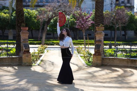 Foto de Hermosa mujer de pelo rizado largo, bailando flamenco con abanico rojo, se encuentra en Sevilla, España. Lleva falda larga negra, camisa blanca y hace diferentes poses de baile. Patrimonio cultural de la humanidad - Imagen libre de derechos
