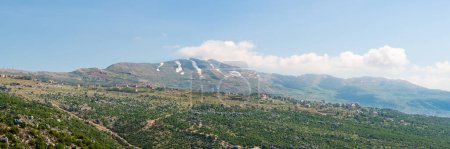 Téléchargez les photos : Un village se trouve sur une crête de colline à travers un paysage rocheux, avec la chaîne de montagnes du Mont Liban, enneigée au printemps, une destination de randonnée populaire au Liban, Moyen-Orient - en image libre de droit