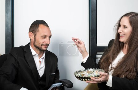 Israelin mit einem schönen Lächeln hält Teller mit Süßigkeiten in der Hand. Lächelnde Frau, die einem jüdischen Geschäftsmann im Amt Süßigkeiten anbietet. Eleganter bärtiger Jude im Yarmulke-Look gibt mir Bonbons