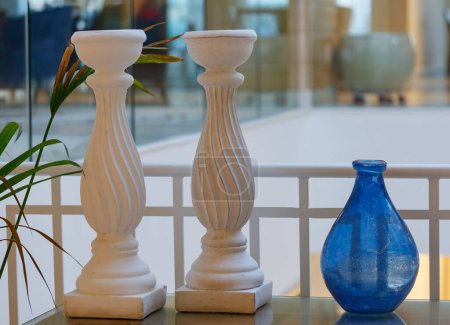 Téléchargez les photos : Vase bleu simple, deux bougeoirs blancs se tiennent sur la table dans le cadre de la décoration de table, décor. Grands chandeliers décoratifs sur un support sur le fond du mur blanc et de la plante verte - en image libre de droit