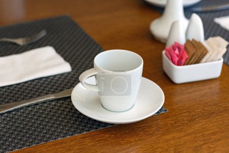 A white coffee cup rests on a saucer, accompanied by cutlery wrapped in a napkin. A dark woven placemat lies underneath the cup, a white holder containing various colored sugar packets. dining table