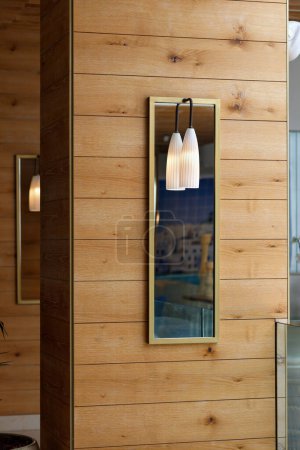 A wooden wall with a long rectangular mirror  embedded into it. Above the mirror, there is a pair of elegant pendant lights with white lampshades that illuminate the space. The interior of the room.