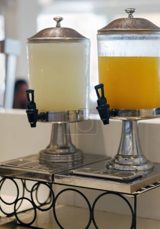 In the dining room two large beverage dispensers stand on a sturdy metal stand. One dispenser holds refreshing lemonade, the other orange juice. Its plastic spigot awaits thirsty patrons. Ice drink. 