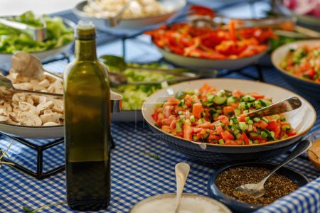 Assortment of fresh salads displayed on various bowls and plates. In the foreground green bottle olive oil. A bowl contains a bright red and green salad made of diced tomatoes and cucumbers, mushrooms