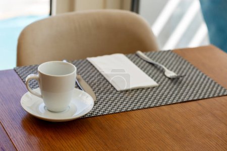 Una taza blanca y un platillo se sientan en un mantel tejido sobre una mesa de madera. Junto a ellos, una servilleta blanca doblada y un tenedor de plata están perfectamente colocados. La silla en el fondo y la luz natural a través de la ventana