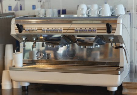The stainless steel professional coffee machine, equipped with multiple options for brewing various types of coffee. White cups are arranged on top, and disposable cups are visible below the spouts