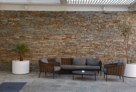 A contemporary outdoor seating area against a textured stone wall. A grey sofa and two chairs with wooden frames are arranged around a black coffee table. A potted plant adds a touch of greenery
