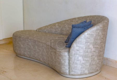 Téléchargez les photos : L'image un canapé moderne avec une forme incurvée unique. Il est tapissé dans un tissu de couleur claire qui a une texture distincte. Un coussin bleu repose sur le canapé, ajoutant une touche de couleur. Le canapé est placé contre un mur uni, et le sol est carrelé - en image libre de droit