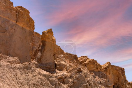 Las formaciones rocosas del paisaje son irregulares y erosionadas, lo que indica cambios geológicos a lo largo del tiempo. El cielo está adornado con suaves nubes iluminadas en tonos de rosa y azul, creando una atmósfera etérea. Mar Muerto, Israel