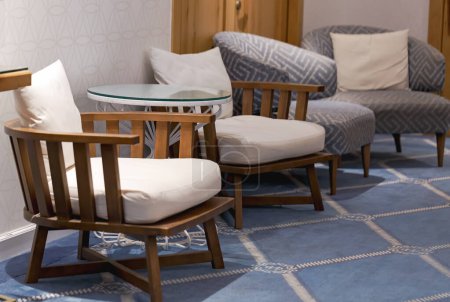 Gemütliches Wohnzimmer mit zwei Holzstühlen mit weißen Kissen und einem Glastisch. Der Boden ist mit einem blauen Teppich verziert, der ein kompliziertes Design aufweist. Die Mauer im Hintergrund. Modernes Zuhause