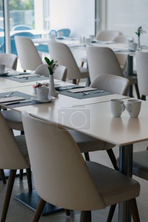 White tables with striped placemats and white cups are paired with chairs. Small vases holding green plants adorn the tables. Large windows allow natural light to illuminate the space. Modern interior