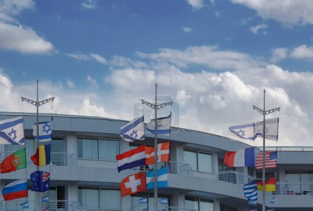 Drapeaux internationaux sur un bâtiment moderne sous un ciel partiellement nuageux. Les drapeaux représentent différents pays, mettant en valeur l'unité mondiale. Les drapeaux sont montés sur des poteaux fixés au bâtiment. Concept de politique