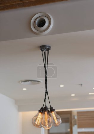 Lustre moderne avec plusieurs ampoules. Les ampoules sont éclairées et suspendues au plafond par des fils noirs. Au-dessus du lustre, une ventilation circulaire est visible. Le plafond est blanc. L'ampoule d'Edison s'allume à partir du courant électrique