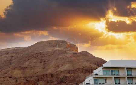 Mountain Sodom surface revealing layers of stone salt under sun rays pierce through dark clouds casting. Building with multiple balconies is juxtaposed against the natural backdrop near the Dead Sea