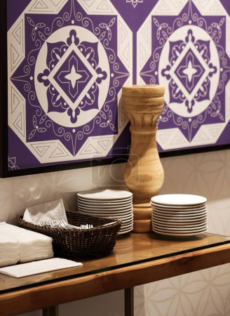 Stapelweise weiße Teller werden vor dem Hintergrund einer violetten Wand mit weißen Mustern verteilt. Esstisch mit Holztisch verziert mit weißen Tellern Servietten, Besteck, Vase. Geschirr gestapelt