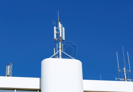 Ein weißes Gebäude steht unter einem strahlend blauen Himmel, auf dem Dach komplexe metallische Antennen und Kommunikationseinrichtungen. Die fortschrittliche Technologie, die für Sende- oder Kommunikationszwecke verwendet wird.