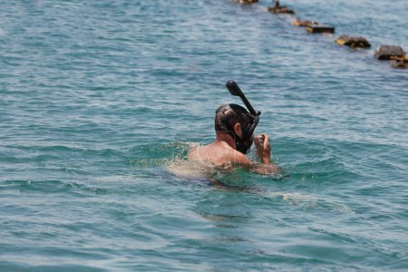 Photo for A man snorkeling in clear blue sea or ocean, adjusting his face mask diving. A black snorkel is clearly seen extending above the guy head. In the background rocks that protrude from the water surface - Royalty Free Image