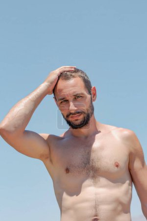 A caucasian strong man standing against a clear blue sky. Handsome bearded male is shirtless looking thoughtfully  at camera. One arm is raised towards the head. Shadows are cast on the persons body