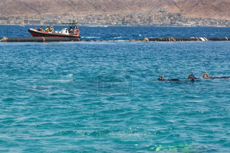 Photo for A boat with people is visible near floating structure. A kids are swimming, enjoy exploring the sea while snorkeling and diving. The backdrop features a distant cityscape nestled against rolling hills - Royalty Free Image