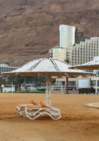 White umbrellas and lounge chairs are prominent in the foreground, placed on sandy ground for relaxation. The background contrasts with a large rugged mountain under a clear sky. Dead Sea, Israel